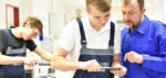 Meisterausbildung Teil IV Ausbildereignung Eine Person in blauer Arbeitskleidung zeigt einer Person in Latzhose die Bearbeitung eines Werkstücks mit einem Handwerkzeug, während im Hintergrund eine weitere Person an einer Werkbank arbeitet.