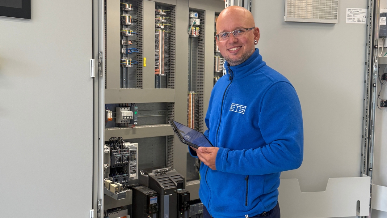 Der Elektrotechnikmeister Dennis Schulz schaut in die Kamera, während er ein Tablet in der Hand hält. Er steht vor einem geöffneten Schaltschrank mit elektrischen Steuerungs- und Schaltkomponenten.