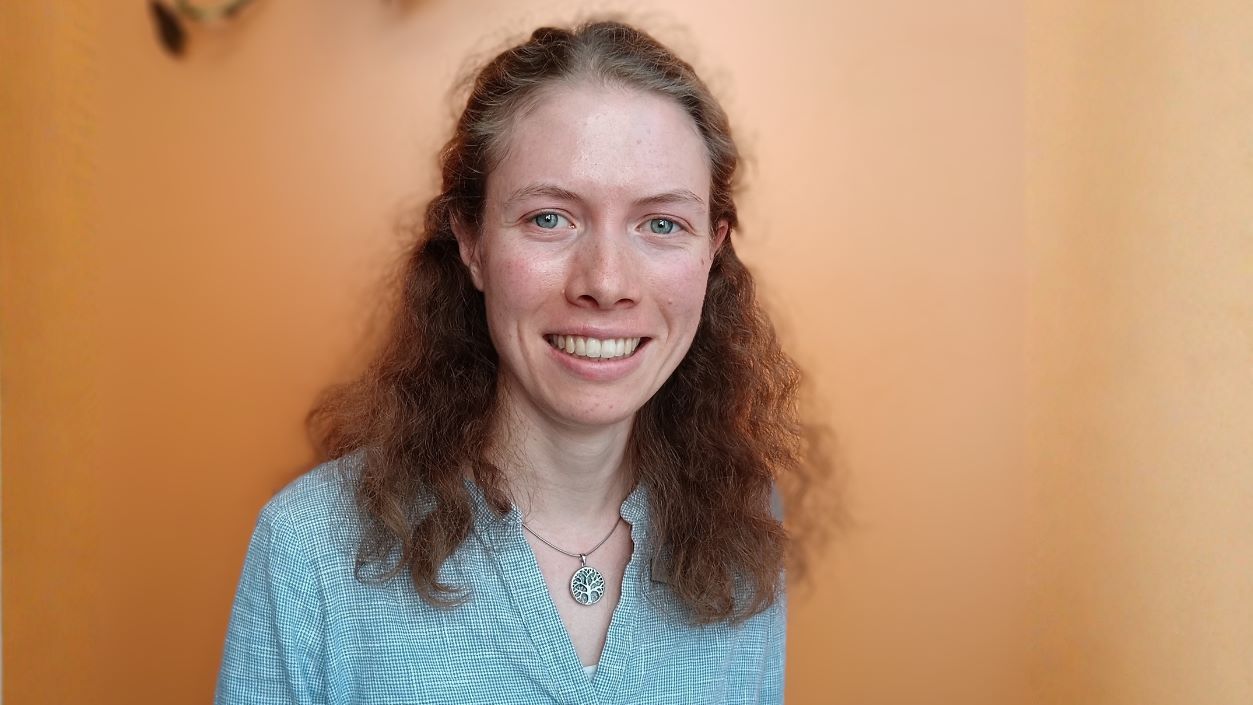 Die Hörakustikermeisterin Ellen Pestel blickt in die Kamera. Sie steht vor einem warmen, orangenen Hintergrund und trägt eine hellblaue Bluse sowie eine Kette mit rundem Anhänger auf dem ein Baum zu sehen ist.