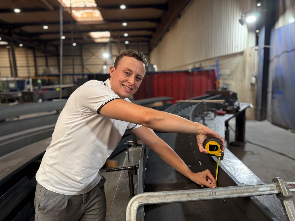Der Metallbaumeister Leon Thiele schaut in die Kamera, wärend er mit einem gelben Maßband ein großes Stahlbauteil in einer Werkhalle misst.