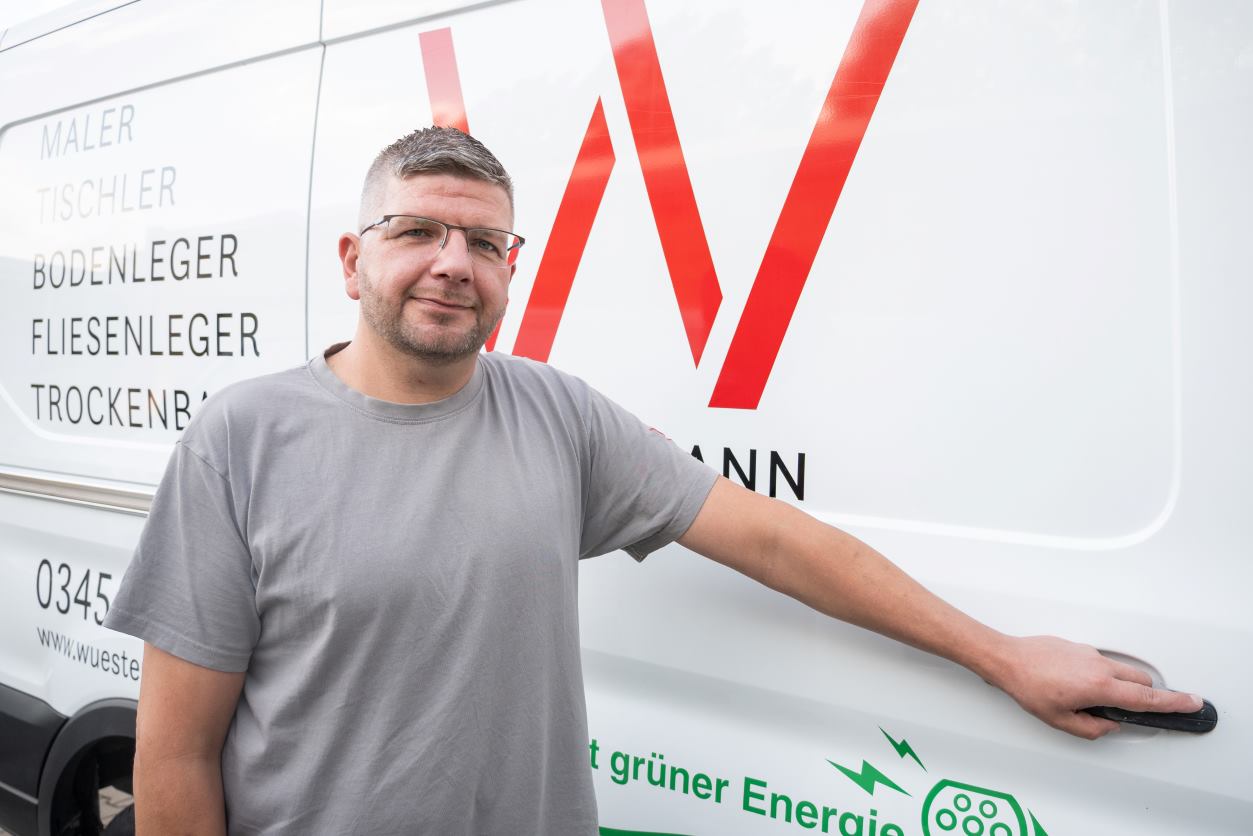 Der ausgezeichnete Handwerksmeister Marcel Kaiser schaut in die Kamera während er vor dem Firmenfahrzeug seines Betriebs steht und die linke Hand an der Schiebetür hält, um sie zu öffnen.