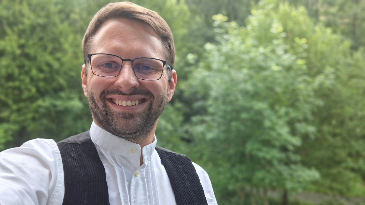 Zimmerermeister Philipp Rittierodt steht vor einem grünen Hintergrund aus Bäumen und blickt in die Kamera. Er trägt eine traditionelle Zimmererkluft.