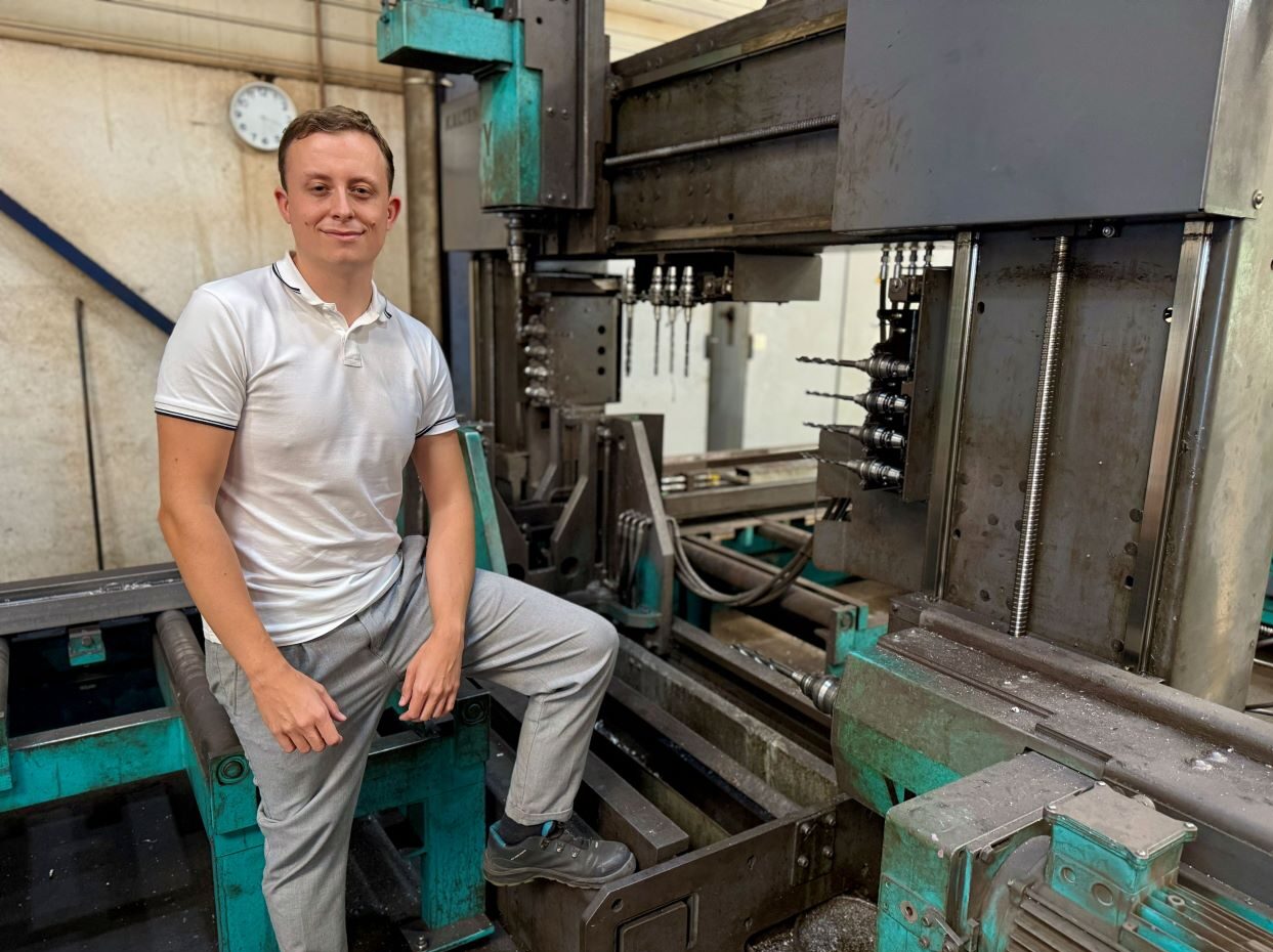 Der Metallbaumeister Leon Thiele schaut in die Kamera während er auf dem Gestell einer großen Metallbearbeitungsmaschine in einer Werkhalle sitzt.