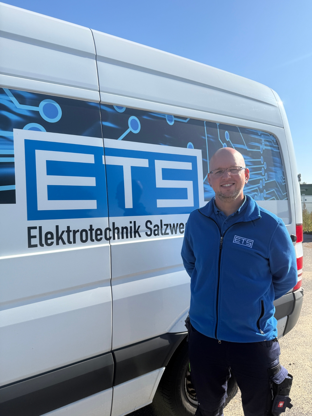 Elektrotechnikermeister Dennis Schulz schaut in die Kamera während er vor dem Firmenfahrzeug seines Betriebes mit dem Schriftzug „ETS Elektrotechnik Salzwedel“ steht.