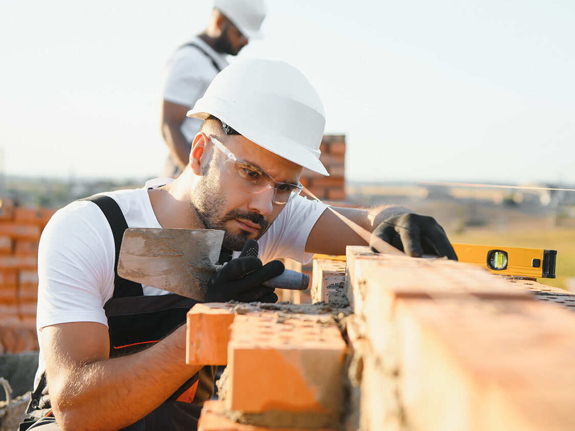 Eine Person mit Schutzhelm, Schutzbrille und Handschuhen setzt auf einer Baustelle eine Ziegelmauer mit einer Kelle; im Hintergrund steht eine weitere Person zwischen gestapelten Ziegeln.