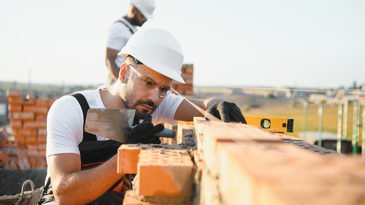 Eine Person mit Schutzhelm, Schutzbrille und Handschuhen setzt auf einer Baustelle eine Ziegelmauer mit einer Kelle; im Hintergrund steht eine weitere Person zwischen gestapelten Ziegeln.