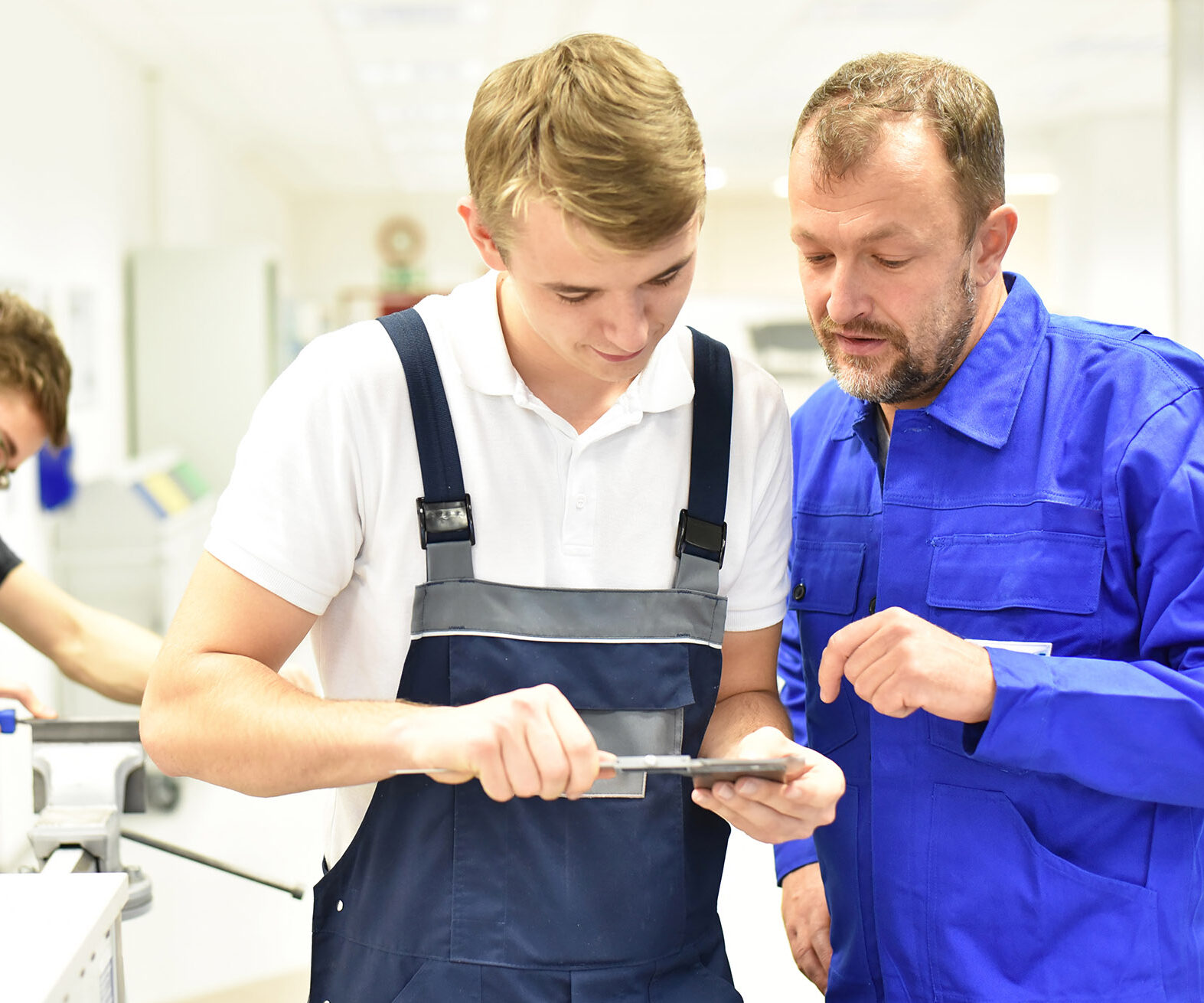 Eine Person in blauer Arbeitskleidung zeigt einer Person in Latzhose die Bearbeitung eines Werkstücks mit einem Handwerkzeug, während im Hintergrund eine weitere Person an einer Werkbank arbeitet.