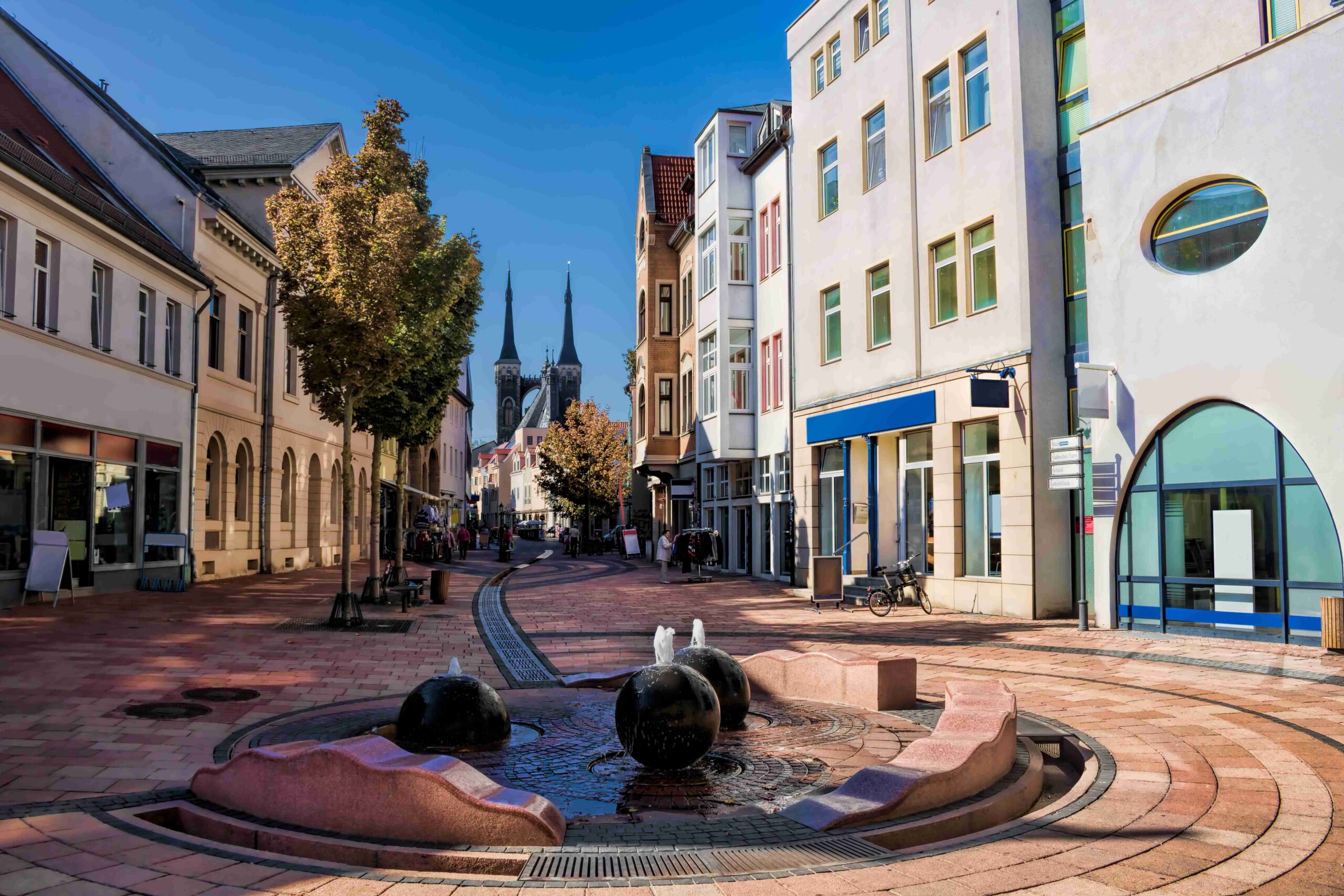Gepflasterte Fußgängerstraße mit einem kleinen Brunnen im Vordergrund, gesäumt von mehrstöckigen Gebäuden und Bäumen; im Hintergrund ragen die Türme einer Kirche unter blauem Himmel auf.