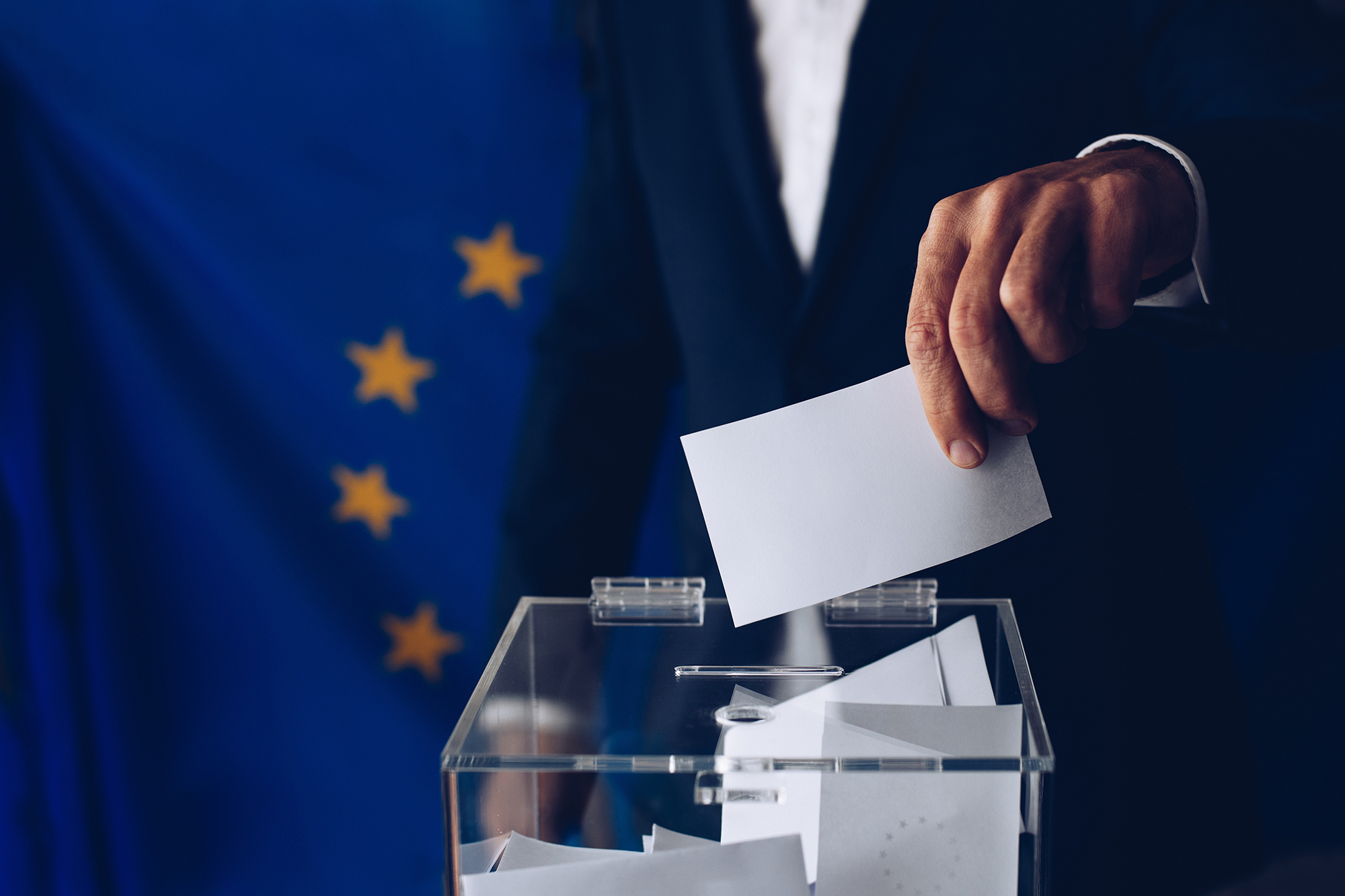 Mann steckt Zettel in Wahlurne, im Hintergrund die Europaflagge