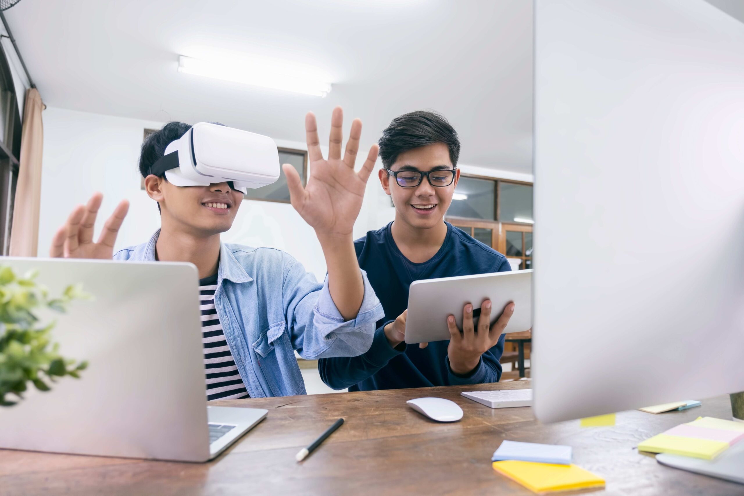 Zwei Personen arbeiten an einem Schreibtisch mit Laptop und Tablet. Eine Person nutzt eine Virtual-Reality-Brille und bewegt die Hände zur Interaktion mit einer virtuellen Anwendung.