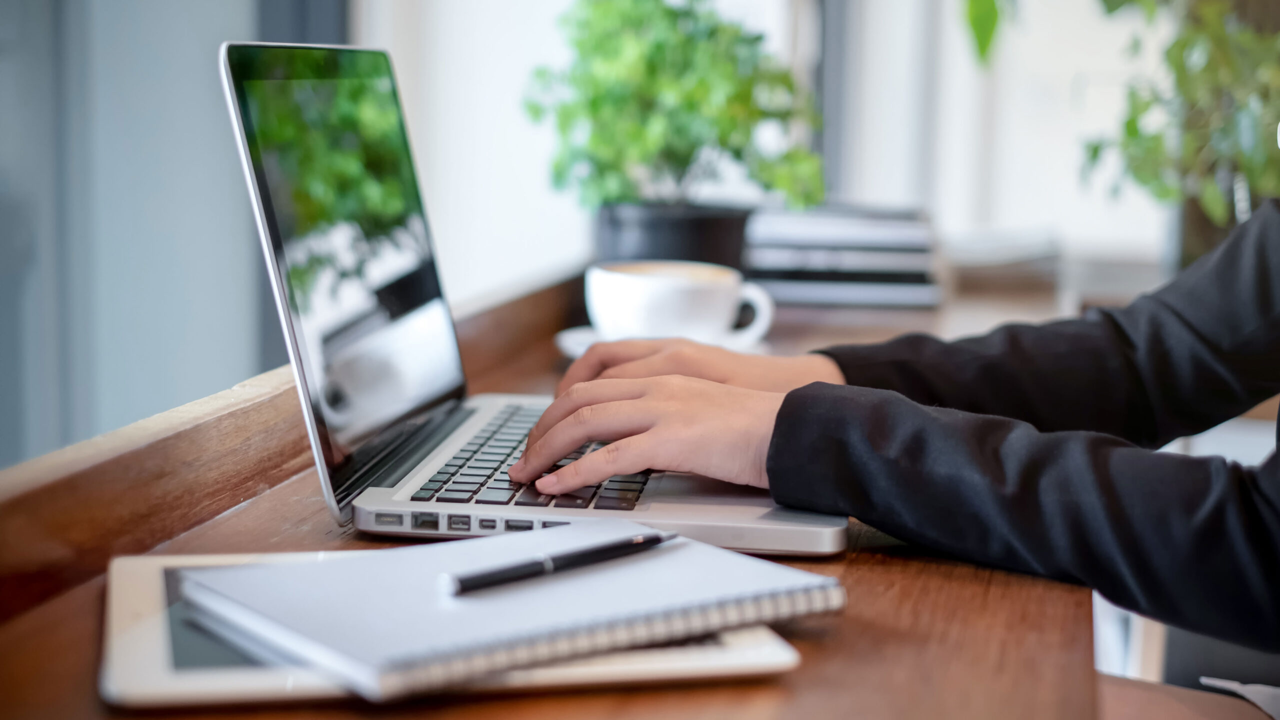 Hände tippen auf einer geöffneten Laptop-Tastatur auf einem Holztisch, daneben liegen ein Notizbuch mit Stift und eine Kaffeetasse, im Hintergrund stehen Pflanzen am Fenster.