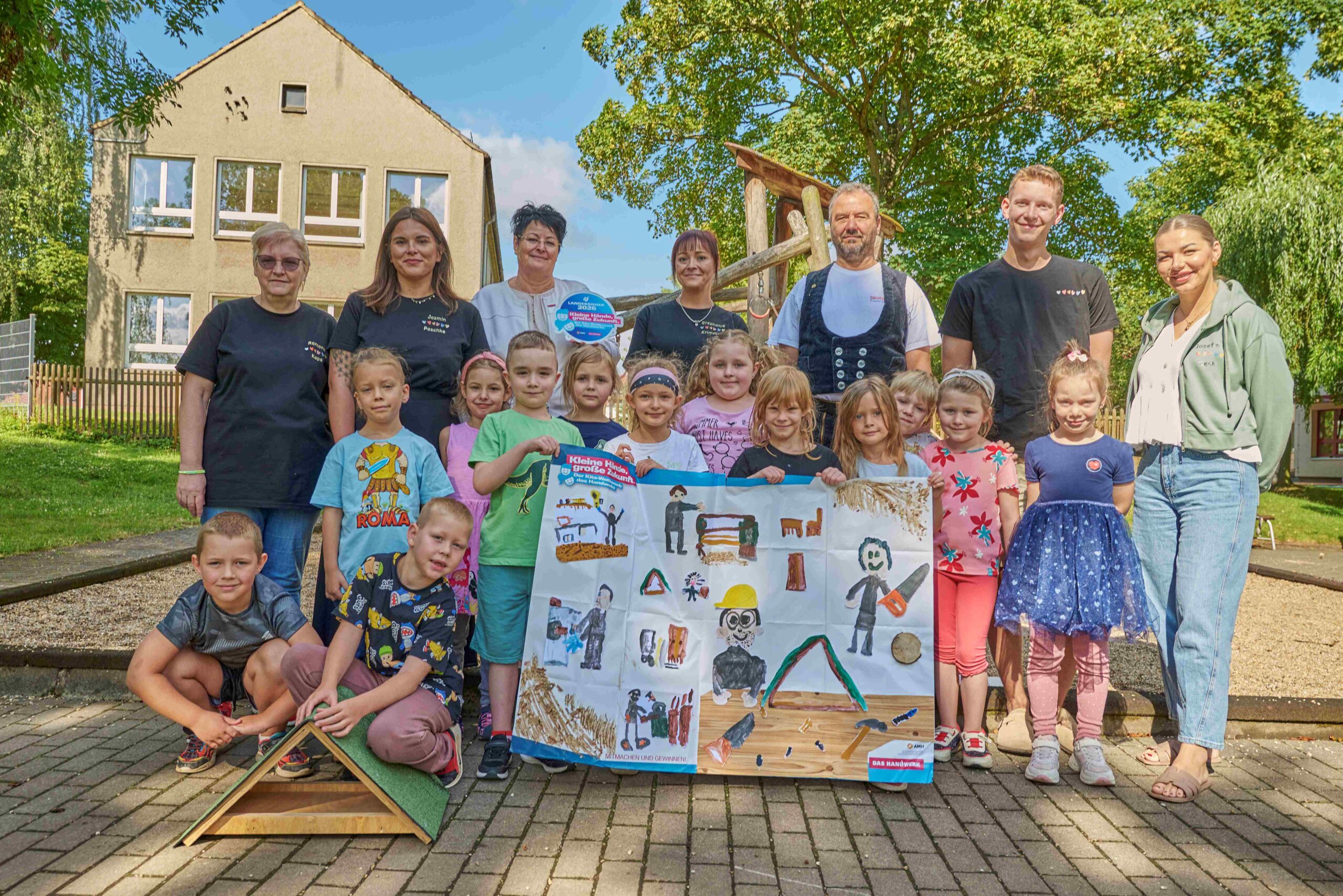 Viele Kitakinder stehen und lächeln in die Kamera. Einige von ihnen halten ein Riesenposter, auf dem gemalte Bauarbeiter bei der Arbeit zu sehen sind. Im Hintergrund sind Frauen und Männer zu sehen, die auch in die Kamera lächeln.