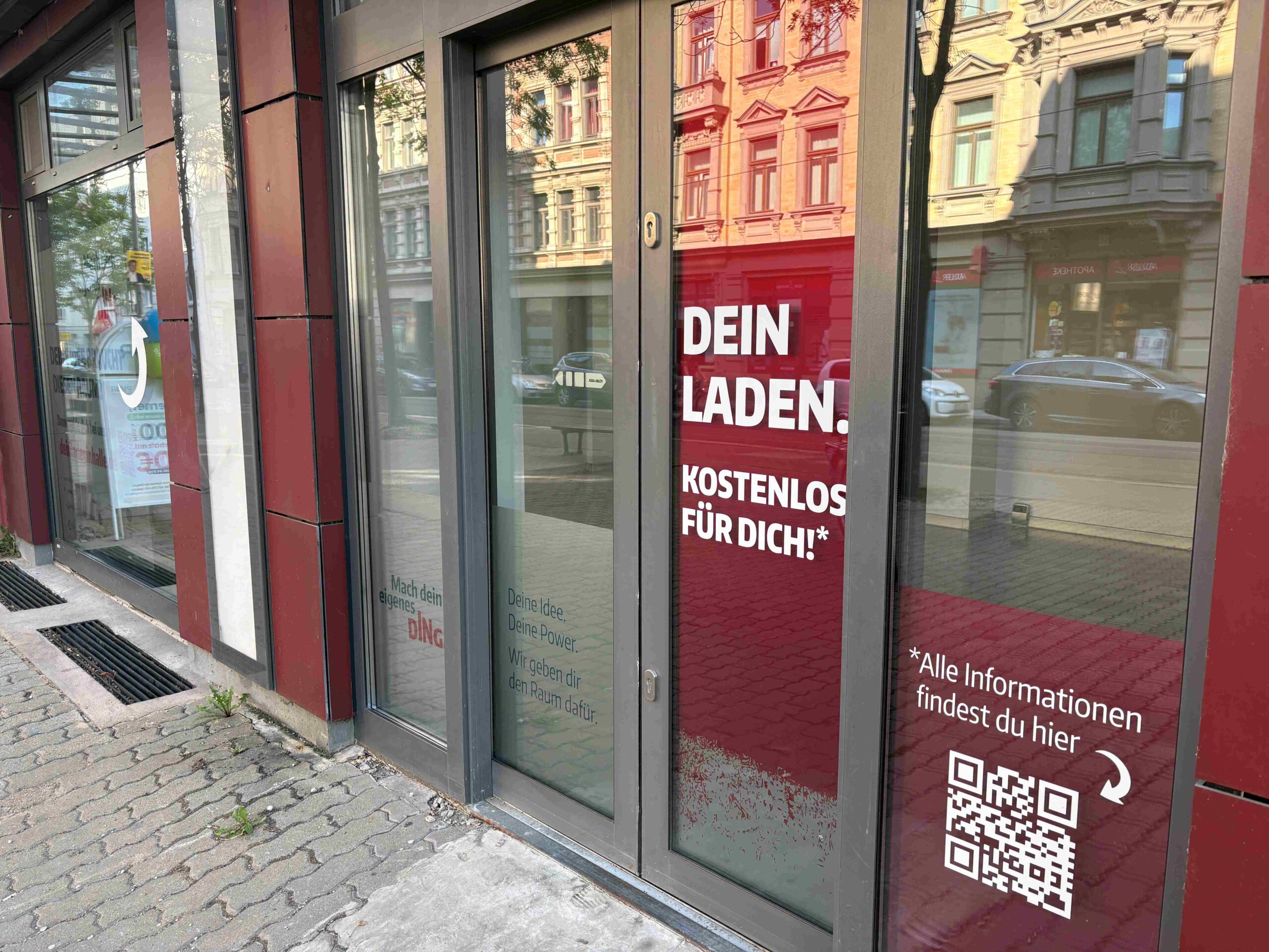 Glasfront einer leerstehenden Ladenfläche mit roter Werbefolie auf der Tür, auf der „DEIN LADEN. KOSTENLOS FÜR DICH!“ sowie ein QR-Code zu sehen sind; in den Scheiben spiegeln sich Gebäude und Fahrzeuge der gegenüberliegenden Straßenseite.