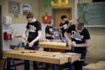 Vier Personen arbeiten in einer Werkstatt an Werkbänken mit Holz, Werkzeugen und einer Maschine; im Hintergrund zeigt eine Person auf eine Tafel mit Skizzen.