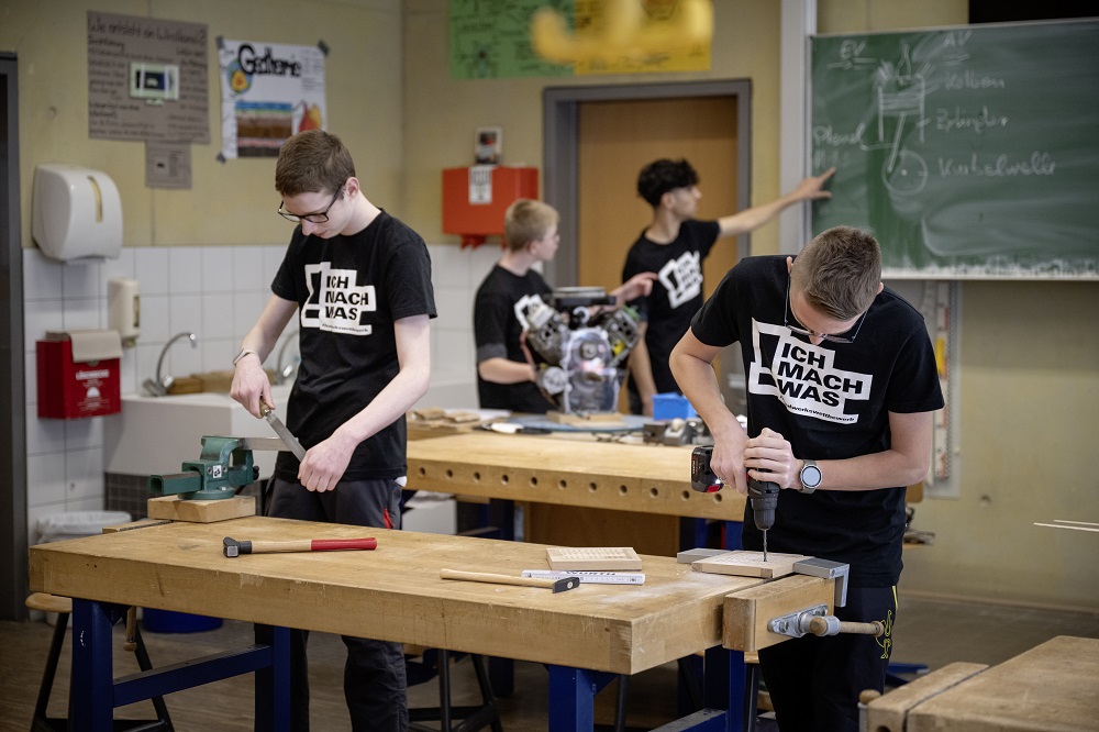 Vier Personen arbeiten in einer Werkstatt an Werkbänken mit Holz, Werkzeugen und einer Maschine; im Hintergrund zeigt eine Person auf eine Tafel mit Skizzen.