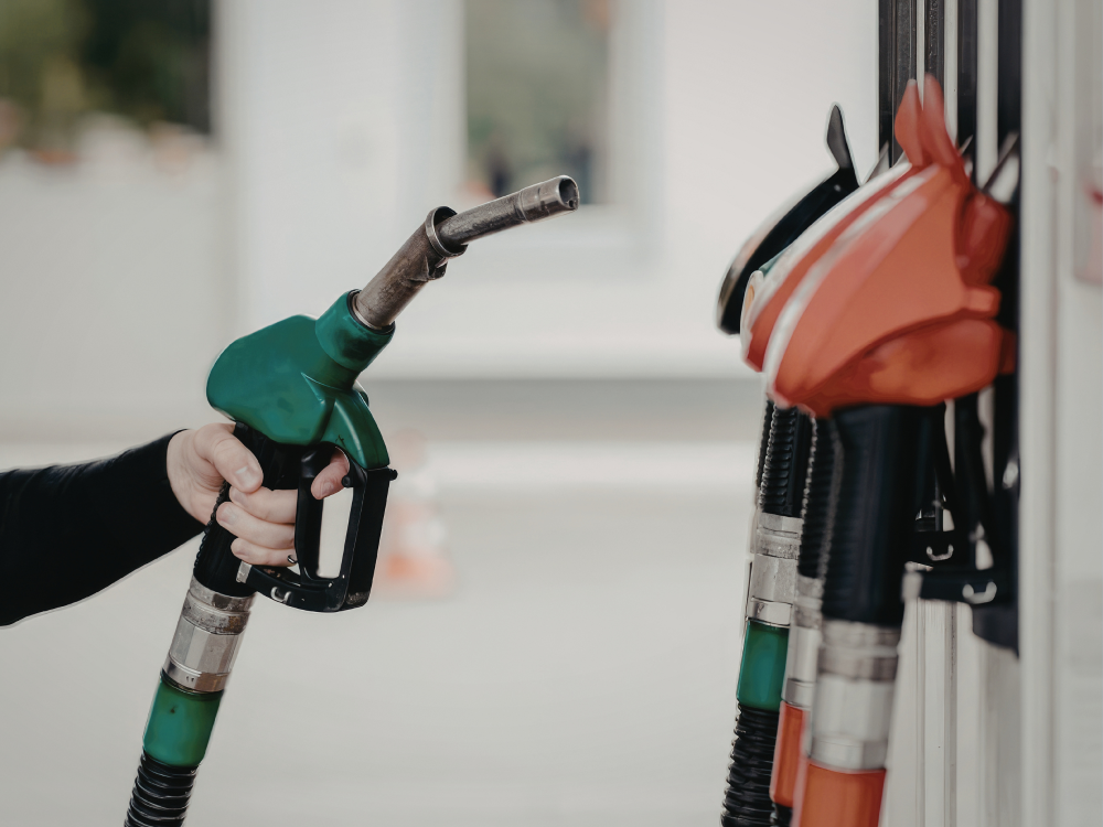 Hand hält eine grüne Zapfpistole an einer Tankstelle; weitere Zapfpistolen hängen daneben.