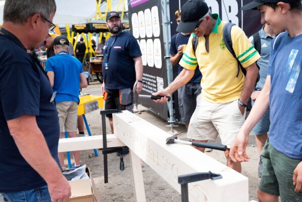 Handwerkskammer Halle Eine Person schlägt mit einem Hammer auf einen eingespannten Holzbalken, während mehrere Besucherinnen und Besucher zusehen.