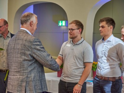 Eine Person im karierten Sakko schüttelt einer Person mit Namensschild die Hand, während weitere Personen in einem festlich gestalteten Innenraum danebenstehen.