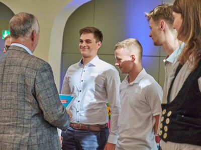 Eine Person im karierten Sakko spricht mit mehreren Personen mit Namensschildern in einem Innenraum mit Rundbögen und dekorativer Beleuchtung.