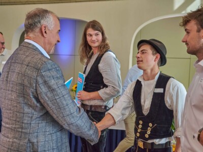 Eine Person im karierten Sakko schüttelt einer Person in traditioneller Arbeitskleidung die Hand, während weitere Personen mit Mappen und Namensschildern danebenstehen.