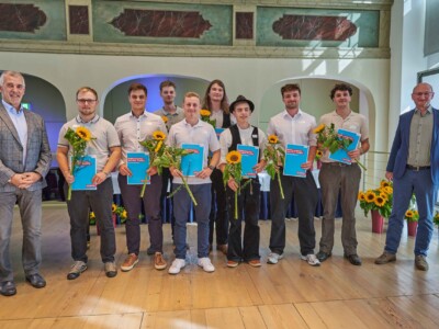 Neun Personen stehen auf einer Bühne mit Sonnenblumen und blauen Mappen für ein Gruppenfoto, flankiert von zwei weiteren Personen am Rand der Bühne.
