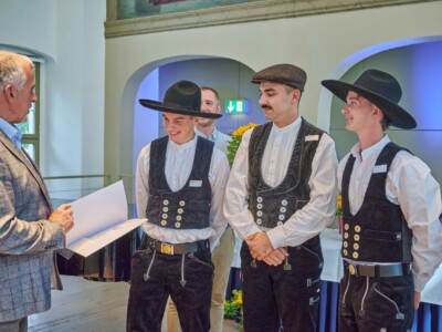 Drei Personen in traditioneller Handwerkskleidung stehen vor einer Person mit geöffneter Mappe in einem festlich gestalteten Innenraum.