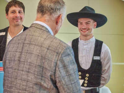 Eine Person in traditioneller Handwerkskleidung mit breitkrempigem Hut steht einer Person im karierten Sakko gegenüber, während eine weitere Person mit Mappe im Hintergrund zu sehen ist.