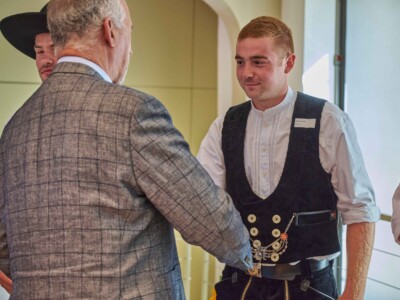 Eine Person in traditioneller Handwerkskleidung schüttelt einer Person im karierten Sakko die Hand.