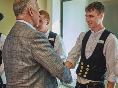 Zwei Personen in traditioneller Handwerkskleidung begrüßen nacheinander eine Person im karierten Sakko mit Handschlag.