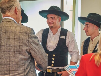 Eine Person in traditioneller Handwerkskleidung mit breitkrempigem Hut begrüßt eine Person im karierten Sakko, während weitere Personen danebenstehen.