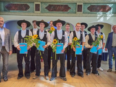 Mehrere Personen in traditioneller Handwerkskleidung stehen mit Sonnenblumen und blauen Mappen für ein Gruppenfoto auf einer Bühne, flankiert von zwei weiteren Personen.