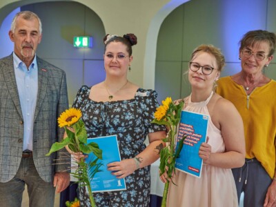 Zwei Frauen mit Sonnenblumen und blauen Mappen stehen zwischen zwei weiteren Personen für ein Gruppenfoto in einem Saal.