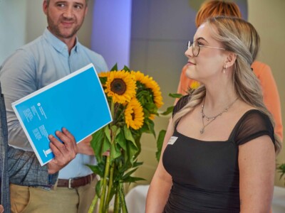 Eine Frau mit blauer Mappe spricht mit einer Person in schwarzem Kleid, während im Hintergrund Sonnenblumen und weitere Personen zu sehen sind.