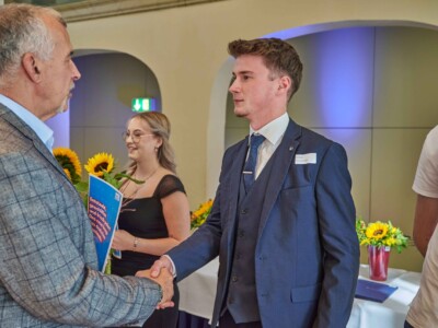 Ein Mann im blauen Anzug schüttelt einer Person im karierten Sakko die Hand; im Hintergrund steht eine Person mit Sonnenblumen und Mappe.