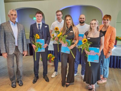 Sieben Personen stehen für ein Gruppenfoto in einem Saal, mehrere halten Sonnenblumen und blaue Mappen.