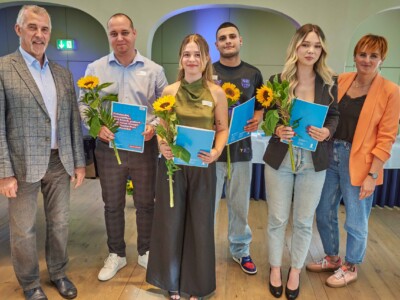 Fünf Personen mit Sonnenblumen und blauen Mappen stehen zwischen zwei weiteren Personen für ein Gruppenfoto in einem Saal.