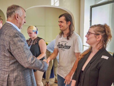 Eine ältere Person im karierten Sakko begrüßt bei einer Veranstaltung einen jungen Erwachsenen per Handschlag, während weitere Teilnehmende in einer Reihe warten.