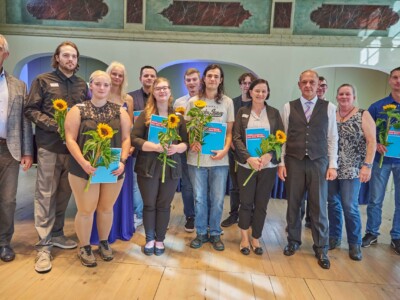 Gruppenfoto mehrerer Teilnehmender einer Feierstunde mit Sonnenblumen und blauen Mappen.