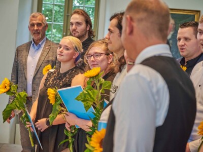 Gruppenfoto mehrerer Teilnehmender einer Feierstunde mit Sonnenblumen und blauen Mappen.