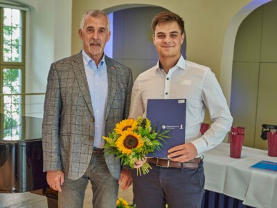 Zwei Personen posieren mit Blumenstrauß und Urkundenmappe für ein Erinnerungsfoto.
