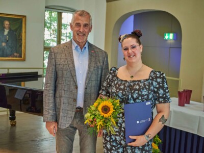 Ein Mann im karierten Sakko steht neben einer Frau mit Blumenstrauß und blauer Mappe der Handwerkskammer Halle (Saale). Beide posieren für ein Erinnerungsfoto in einem Veranstaltungsraum.