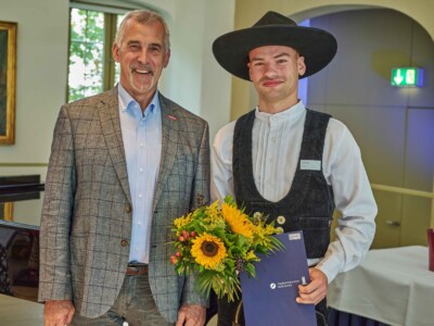 Ein Mann im karierten Sakko steht neben einem jungen Mann in traditioneller Handwerkskleidung mit breitkrempigem Hut. Der junge Mann hält einen Blumenstrauß mit Sonnenblumen und eine Urkundenmappe der Handwerkskammer Halle (Saale).