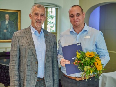 Ein Mann im karierten Sakko und ein Mann im hellblauen Hemd stehen nebeneinander für ein Erinnerungsfoto. Der Absolvent hält einen Blumenstrauß mit Sonnenblumen sowie eine blaue Urkundenmappe der Handwerkskammer Halle (Saale).