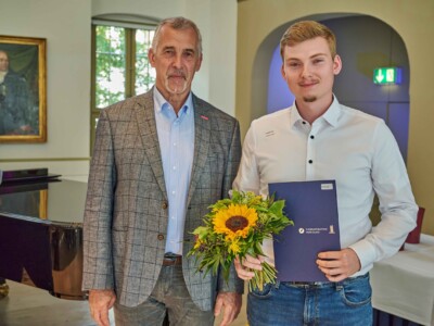 Ein Mann im weißen Hemd präsentiert einen Blumenstrauß und eine blaue Urkundenmappe der Handwerkskammer Halle (Saale), während er neben einem Mann im karierten Sakko für ein Erinnerungsfoto steht.