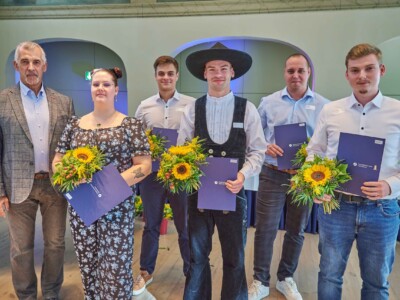 Fünf Absolventinnen und Absolventen stehen mit Blumensträußen und Urkundenmappen der Handwerkskammer Halle (Saale) auf einer Bühne. Links im Bild steht ein Mann im karierten Sakko.