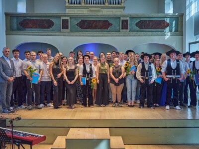 Gruppenfoto einer Abschluss- beziehungsweise Freisprechungsfeier: Rund dreißig Absolventinnen und Absolventen sowie Gäste stehen auf einer Bühne. Mehrere Personen halten Sonnenblumen und Urkundenmappen der Handwerkskammer Halle (Saale).
