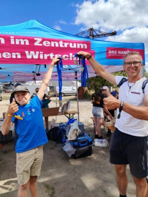 Handwerkskammer Halle Zwei Personen halten vor einem Handwerkszelt Medaillen an blauen Bändern hoch und zeigen jeweils einen erhobenen Daumen.