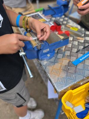 Handwerkskammer Halle Hände feilen eine kleine Metallplatte in einem blauen Schraubstock auf einer Werkbank mit Werkzeugen.