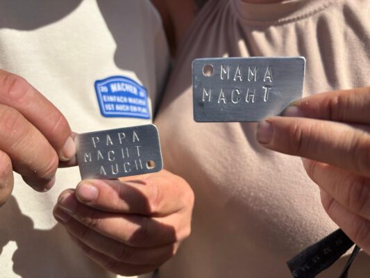 Handwerkskammer Halle Zwei Hände halten kleine gravierte Metallanhänger mit den Aufschriften „PAPA MACHT AUCH“ und „MAMA MACHT“.