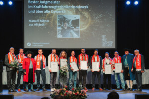 Eine Gruppe von Personen steht auf einer Bühne, mehrere davon halten gerahmte Urkunden und Blumensträuße; einige tragen rote Schals mit der Aufschrift „HANDWERKSMEISTER“, zwei Personen historische Kleidung.